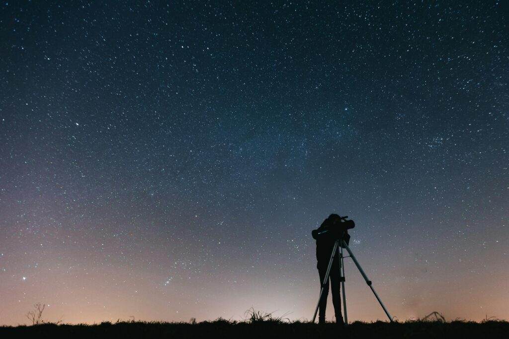 Silhouette of a person looking through a telescope on a tripod under a vast, star-filled night sky.